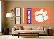 Load image into Gallery viewer, Orange 3x5 foot wide Clemson University flag with Tigers Alumni Logo and two metal grommets covering the tan wall of a well-organized room with couch and pillow