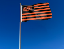 Load image into Gallery viewer, Oklahoma State University Flag with seven black stripes six orange stripes and two metal grommets attached to flagpole waving in the wind