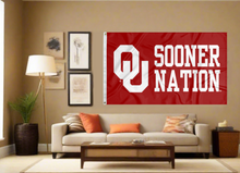 Load image into Gallery viewer, red 3x5 feet University of Oklahoma Flag with OU Sooner Nation and two metal grommets for indoor home decor covering the tan wall of well lit room