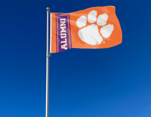 Load image into Gallery viewer, Orange 3x5 feet wide Clemson University Alumni Flag with two metal grommets attached to flagpole waving in the wind