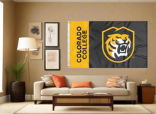 Load image into Gallery viewer, 3x5 foot wide Colorado College Flag with gold stripe and black stripe tiger head shield logo and two metal grommets covering the tan wall of the living room