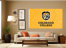 Load image into Gallery viewer, Gold 3x5 foot wide Colorado College Flag with Colorado College logo Tiger head logo and two metal grommets covering the tan wall of a living room