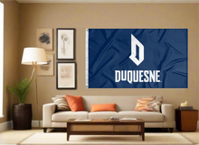 Load image into Gallery viewer, Blue 3x5 foot wide Duquesne University Flag with D Duquesne logo and two metal grommets covering the tan wall of the living room