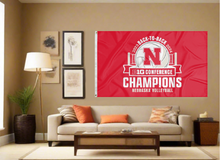 Load image into Gallery viewer, Red 3x5 foot wide Nebraska Big Ten Conference Champion 2024 Flag with Two metal grommets covering a tan wall of well organized room with couch and pillows