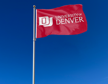 Load image into Gallery viewer, Red 3x5 foot wide University of Denver Flag with white school logo and two metal grommets attached to flagpole waving in the wind