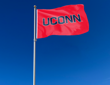 Load image into Gallery viewer, Red 3x5 foot wide University of Connecticut (UCONN) UCONN Red 3x5 Flag with blue UCONN logo and two metal grommets attached to flagpole waving in the wind