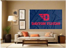 Load image into Gallery viewer, Dark Blue 3x5 foot wide University of Dayton Flyers Blue 3x5 Flag with red D Dayton Flyers logo and two metal grommets covering the tan wall inside of a well organized room with couch pillows and light