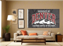 Load image into Gallery viewer, Black 3x5 foot wide University of Denver Flag with Lacrosse Capital of the West Logo and two metal grommets covering the tan wall of a living room with couch and throw pillows
