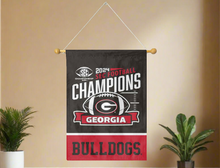 Load image into Gallery viewer, Black and Red 2 panel 30x40 inch long University of Georgia SEC Football Championship 2024 Champions House Flag hanging on a banner pole covering a tan wall with two plants on right and left sides