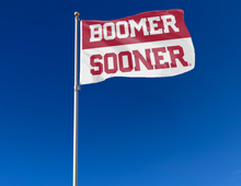Load image into Gallery viewer, Red and White 2 Panel 3x5 University of Oklahoma Flag with Boomer Sooner Logo and two metal grommets for outdoor home decor attached to flagpole waving in the wind