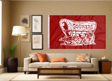 Load image into Gallery viewer, Red 3x5 feet wide Oklahoma Sooners Flag with Sooners Wagon Logo and two metal grommets for indoor home decor covering tan wall of a well lit room