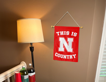 Load image into Gallery viewer, red nebraska garden flag with this is Huskers Country logo hung from banner pole inside the living room
