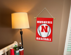 Red 13x18 Nebraska Baseball Garden Flag hung from banner pole inside living room