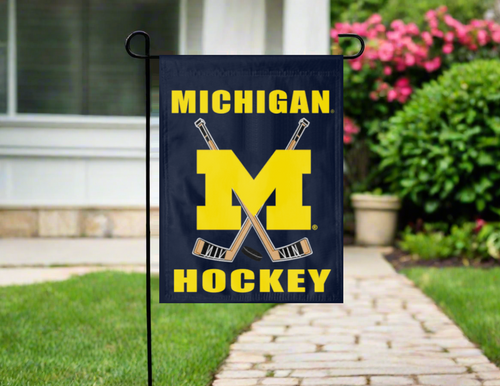 Michigan hockey garden flag with crossed hockey sticks on a blue background, displayed outdoors.