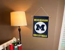 Load image into Gallery viewer, blue Michigan Volleyball garden flag hanging on a banner pole next to a lamp and drinks.
