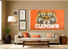 Load image into Gallery viewer, Orange 3x5 foot wide Clemson University Flag with Realistic Tiger Face and Clemson Logos with two metal grommets covering the tan wall inside of a well organized room with couch pillows and light
