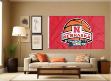 Load image into Gallery viewer, Red 3x5 Nebraska March Madness Flag with two metal grommets covering the tan wall of a well-lit room under couch and coffee table
