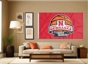 Red 3x5 Nebraska March Madness Flag with two metal grommets covering the tan wall of a well-lit room under couch and coffee table