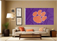 Load image into Gallery viewer, Purple 3x5 feet wide Clemson University Flag with Orange Paw and Two Metal Grommets covering the tan wall inside of a well organized room with couch pillows and light
