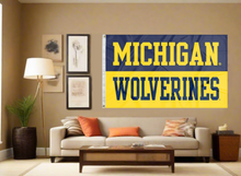Load image into Gallery viewer, 3x5 foot wide University of Michigan Flag with Michigan Wolverines Logo and two metal grommets covering the tan wall of a well-lit room
