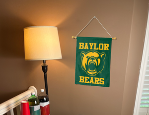 Green 13x18 inch long Baylor University Garden Flag with Sic 'Em Bears Logo hanging on a banner pole covering the well-lit tan wall of the living room