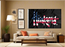 Load image into Gallery viewer, Black 3x5 foot wide University of Nebraska flag with USA flag striped Huskers logo and two metal grommets covering the tan wall of a well-lit room
