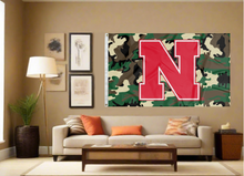 Load image into Gallery viewer, Camo 3x5 University of Nebraska Flag with Red N and two metal grommets covering the tan wall of a well-lit room

