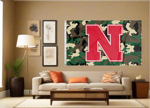 Camo 3x5 University of Nebraska Flag with Red N and two metal grommets covering the tan wall of a well-lit room