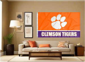 3x5 feet wide Clemson University Tigers orange and Purple stripes with Clemson Tigers Logo and two metal grommets covering the tan wall inside of a well organized room with couch pillows and light
