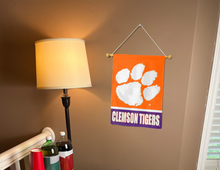 Load image into Gallery viewer, 13x18 inch long Clemson University Garden Flag with orange and Purple stripes and Clemson Tigers Logo hanging on a banner pole covering the tan wall of a well-lit room
