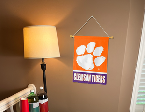 13x18 inch long Clemson University Garden Flag with orange and Purple stripes and Clemson Tigers Logo hanging on a banner pole covering the tan wall of a well-lit room