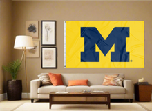 Load image into Gallery viewer, Gold 3x5 foot wide University of Michigan Flag with two metal grommets covering the tan wall of a well-lit room
