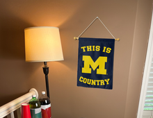 Load image into Gallery viewer, Blue University of Michigan Garden flag with this is Michigan Country logo hung on banner pole next to lamp and drinks
