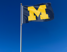 Load image into Gallery viewer, Blue 3x5 foot wide University of Michigan Flag with two metal grommets attached to flagpole waving in the wind
