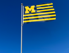 Load image into Gallery viewer, USA Flag style University of Michigan Flag with seven blue stripes six gold stripes attached to flagpole waving in the wind
