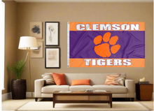Load image into Gallery viewer, 3x5 foot wide three orange purple and orange striped Clemson University Flag with Tigers Logo and two metal grommets covering the tan wall inside of a well organized room with couch pillows and light
