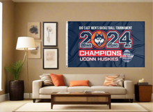 Load image into Gallery viewer, Navy blue 3x5 foot wide UCONN flag with big east men&#39;s basketball tournament 2024 champions uconn huskies logo and two metal grommets covering the tan wall of a well lit room with couch and throw pillows
