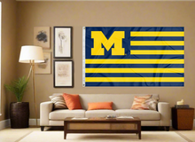 Load image into Gallery viewer, Blue and Gold USA Flag style University of Michigan Flag with seven blue stripes six gold stripes and two metal grommets covering tan wall of a well-lit room
