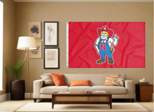 Load image into Gallery viewer, Red University of Nebraska Flag with Herbie Husker Logo covering the tan wall of a well-lit room
