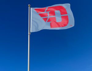 Light blue 3x5 foot wide University of Dayton Flag with red D logo and two metal grommets attached to flagpole waving in the wind
