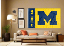 Load image into Gallery viewer, 2 panel 3x5 foot wide University of Michigan with Michigan M Logo and two metal grommets covering the tan wall of a well-lit room
