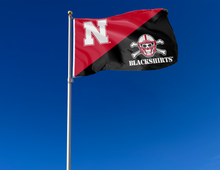Load image into Gallery viewer, 3x5 foot wide red and black University of Nebraska House Divided Flag with N logo and blackshirts logo and two metal grommets attached to flagpole waving in the wind
