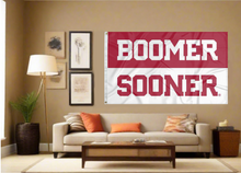 Load image into Gallery viewer, Red and White 2 Panel 3x5 University of Oklahoma Flag with Boomer Sooner Logo and two metal grommets for indoor home decor covering tan wall of well lit room

